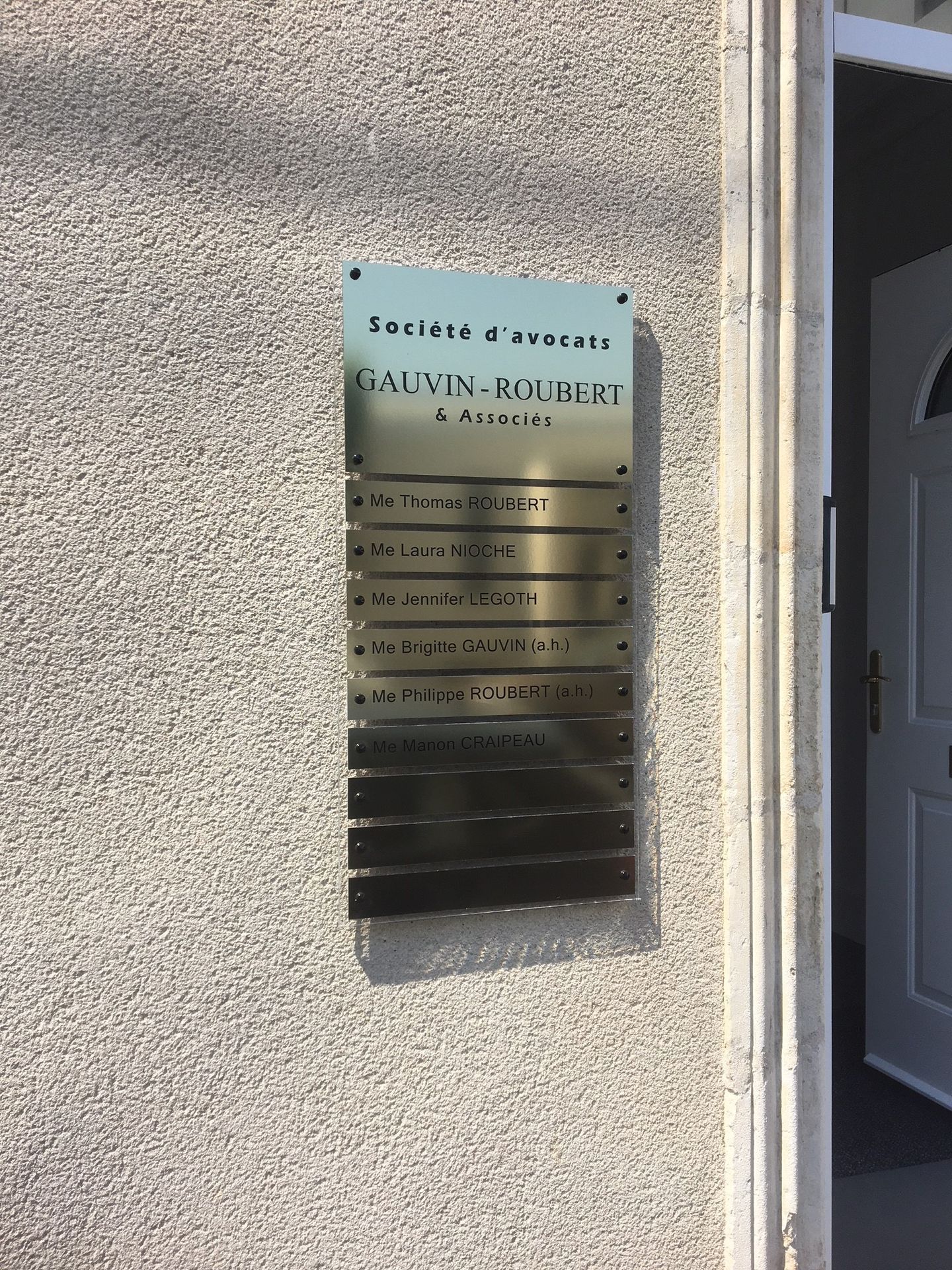 Une enseigne métallique pour immeuble de bureaux, avec du texte sur un mur extérieur texturé gris clair, à côté d'une porte.