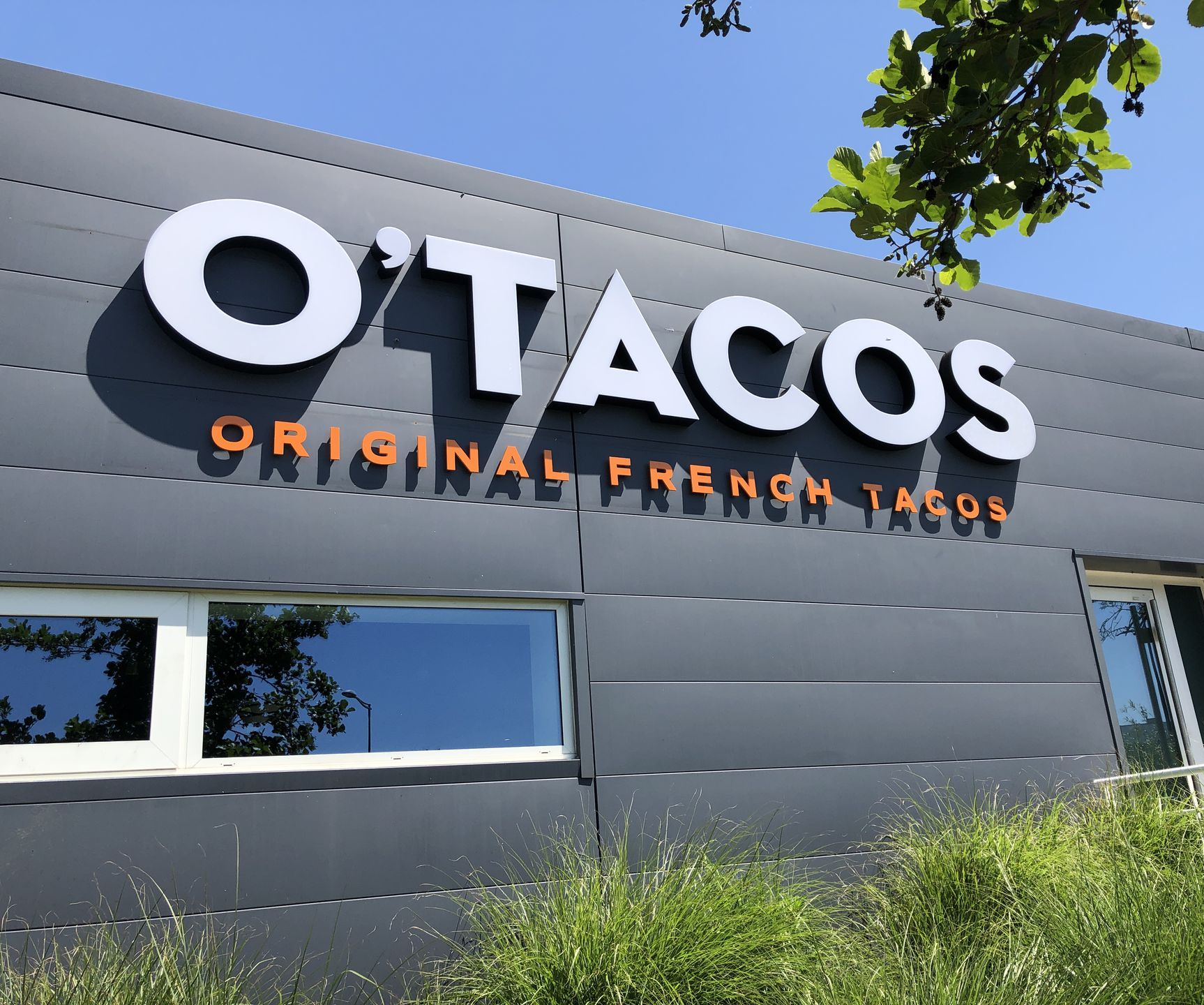L'extérieur d'un restaurant O'Tacos, avec son enseigne blanche à texte orange, se détache sur un bâtiment gris foncé.