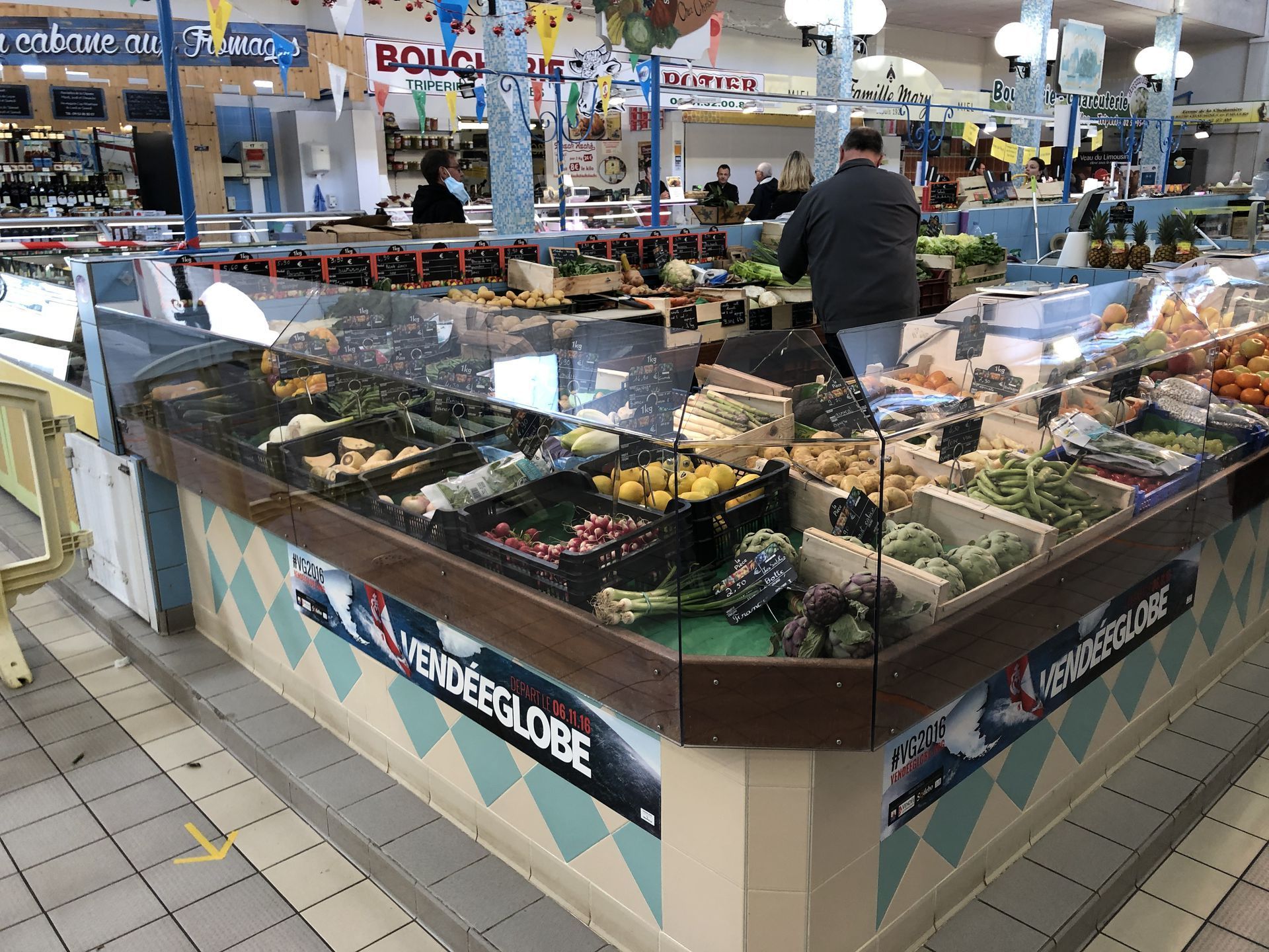 Une personne se tient derrière un grand comptoir d'exposition rempli de produits frais dans un marché couvert aux murs carrelés.