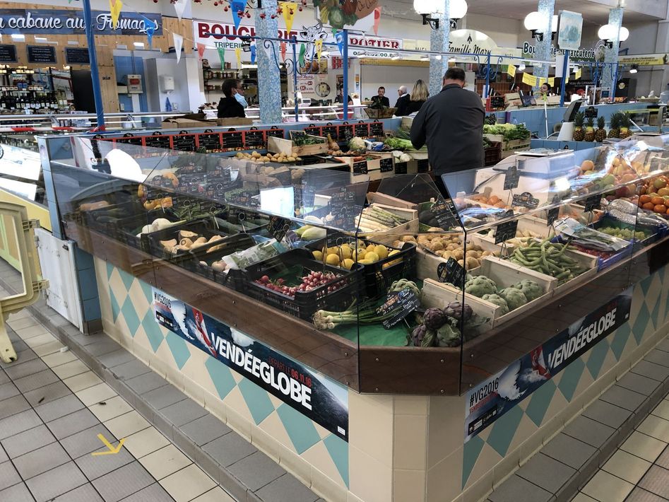 Une vitrine présentant divers fruits et légumes à l'intérieur d'un marché couvert animé, avec un client à proximité.