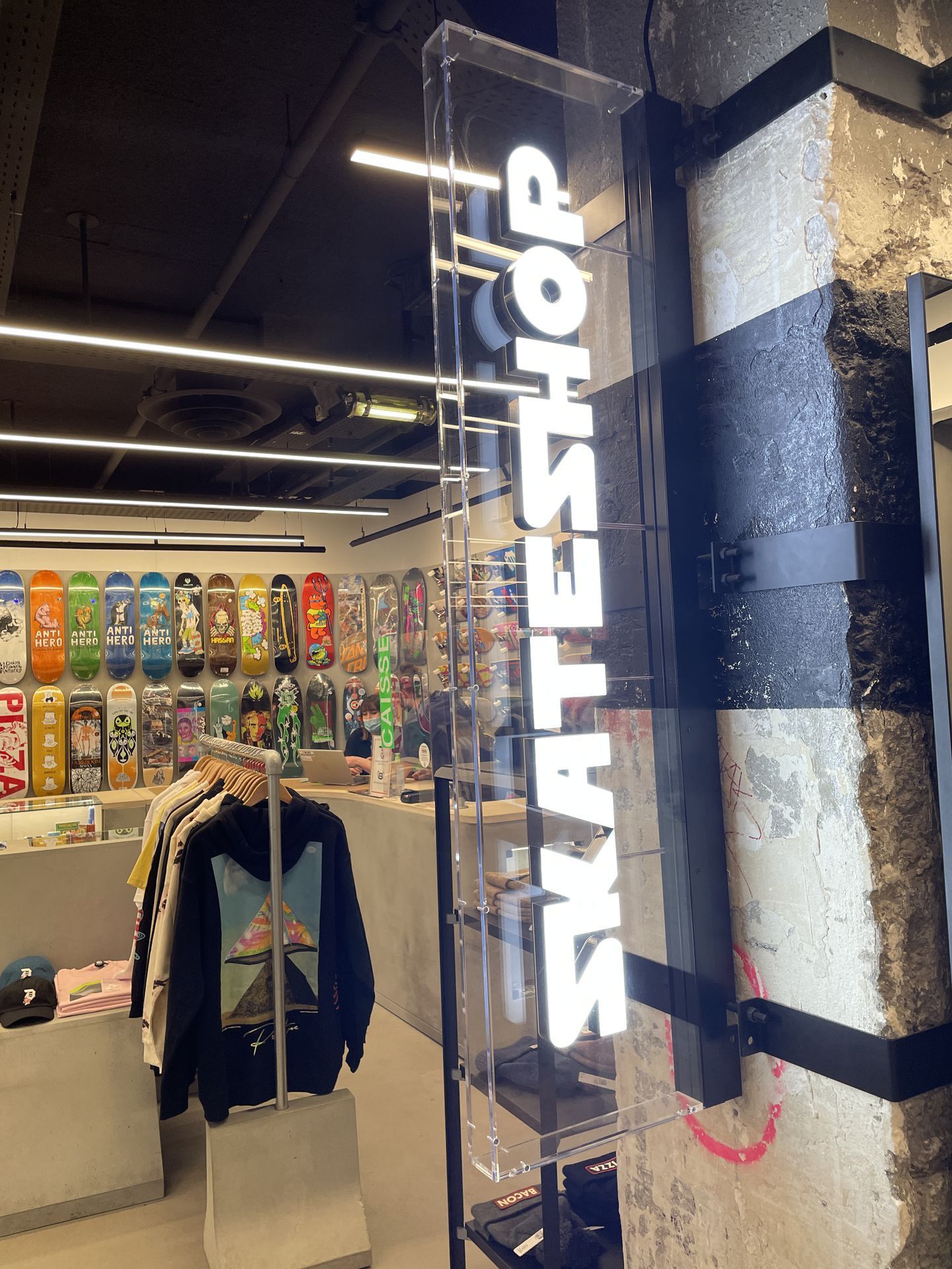 Une enseigne verticale lumineuse SKATESHOP est accrochée dans un magasin présentant des rangées de skateboards colorés et des portants de vêtements.