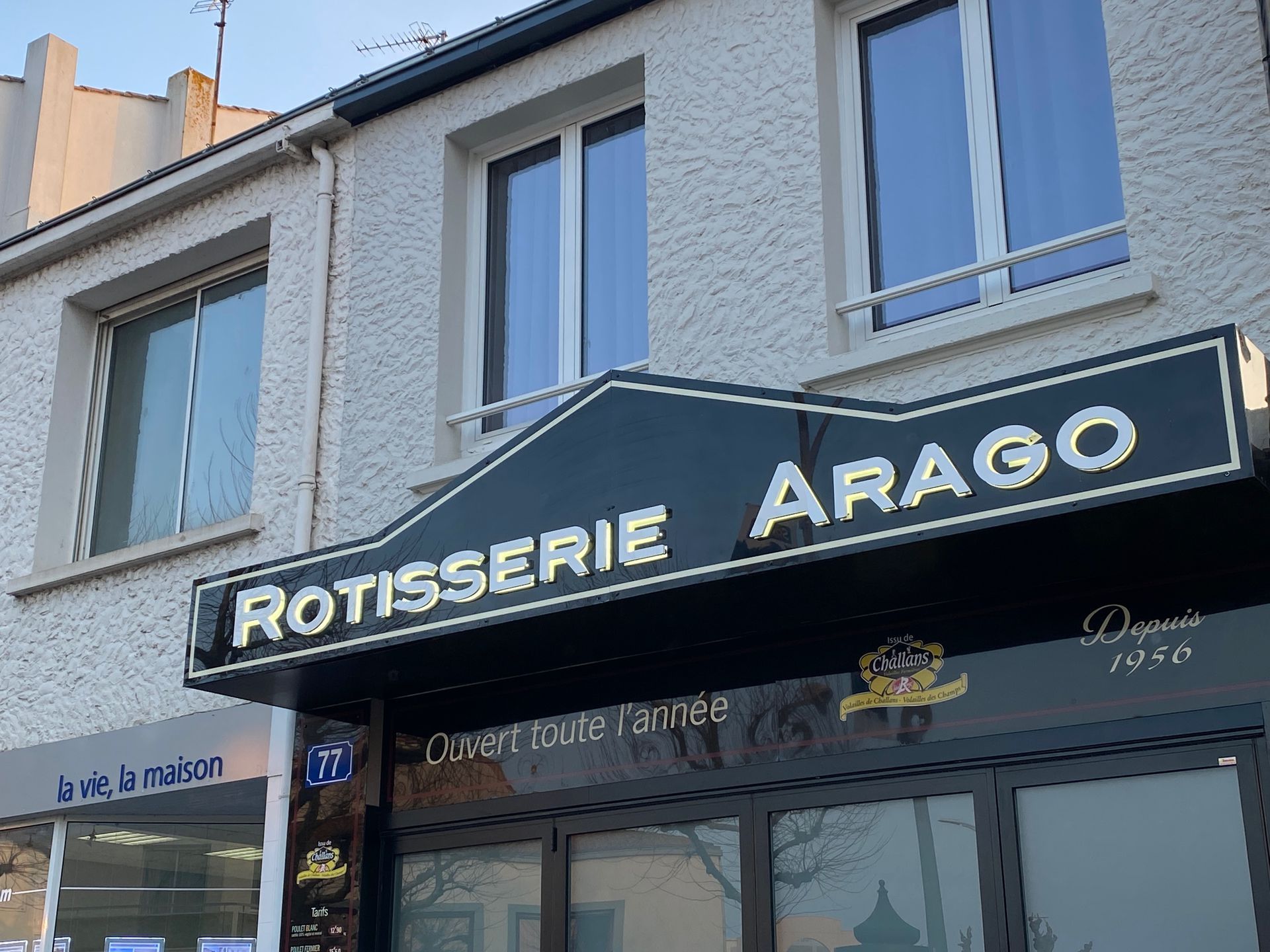 L'extérieur de la rôtisserie Arago, un magasin à l'enseigne noire et aux lettres blanches, situé à un coin de rue sous un ciel bleu.