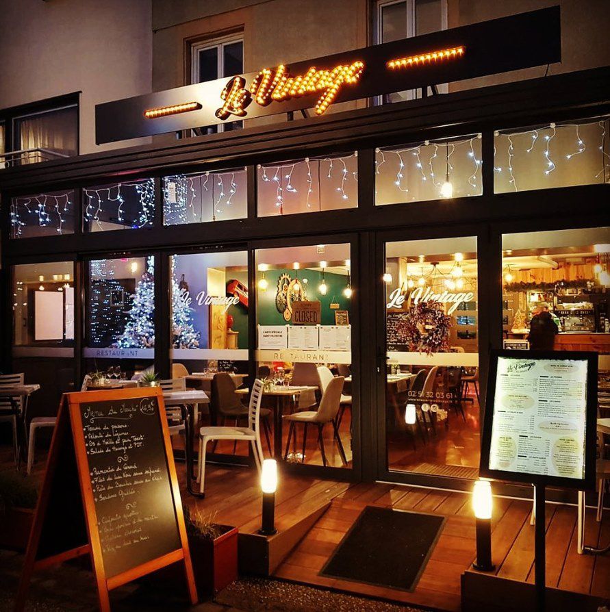 Façade du restaurant la nuit, avec son enseigne lumineuse Le Vintage, ses grandes vitrines et un tableau de menu sur le trottoir.