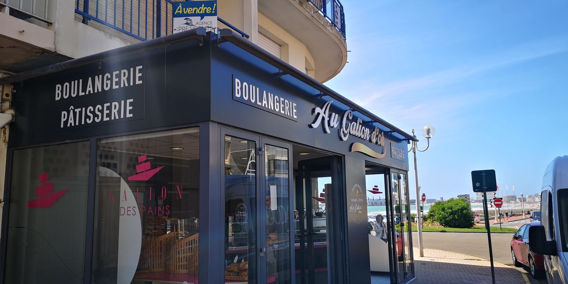 Vue au niveau de la rue d'une boulangerie d'angle avec une enseigne noire indiquant Boulangerie Pâtisserie par une journée ensoleillée.