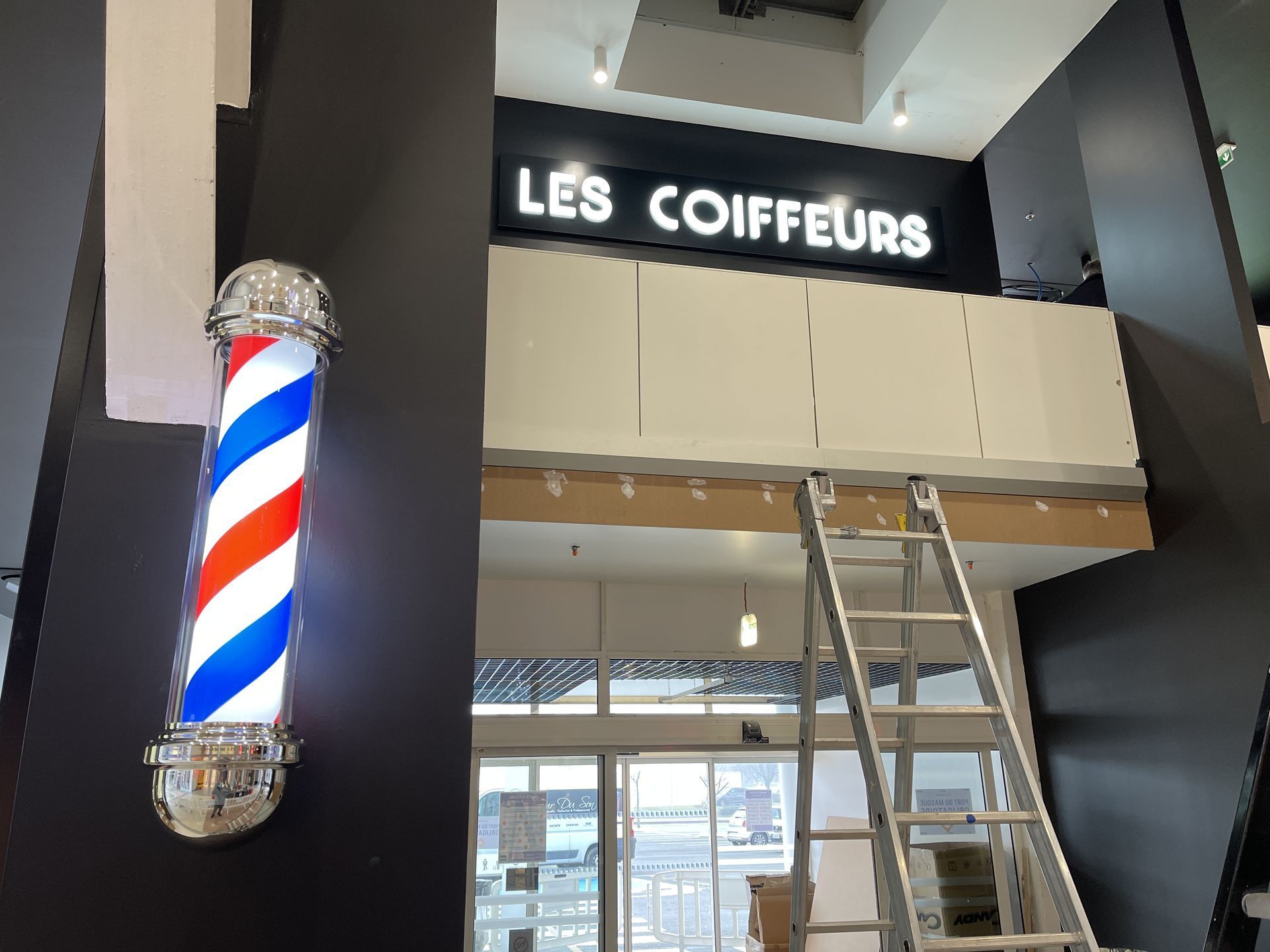 Un poteau de barbier rouge, blanc et bleu vif, fixé sur un mur sombre à côté d'une échelle menant à une mezzanine portant l'inscription LES COIFFEURS.