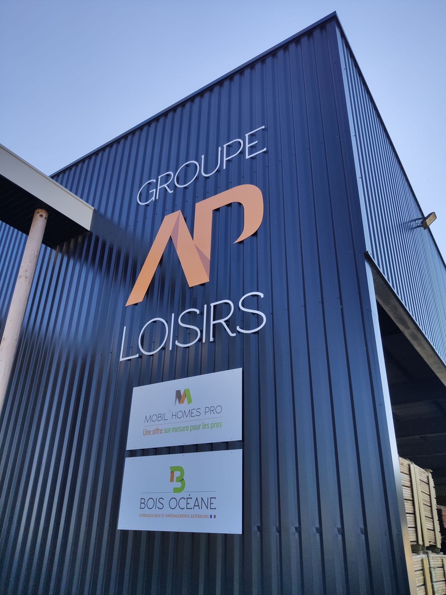 Vue en contre-plongée d'un bâtiment métallique bleu nervuré affichant le logo GROUPE AP LOISIRS et deux enseignes plus petites de l'entreprise.