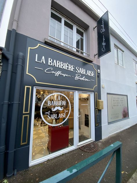 Une devanture avec une enseigne gris foncé, des garnitures dorées et un logo circulaire blanc qui dit La Barbière Sablaise.