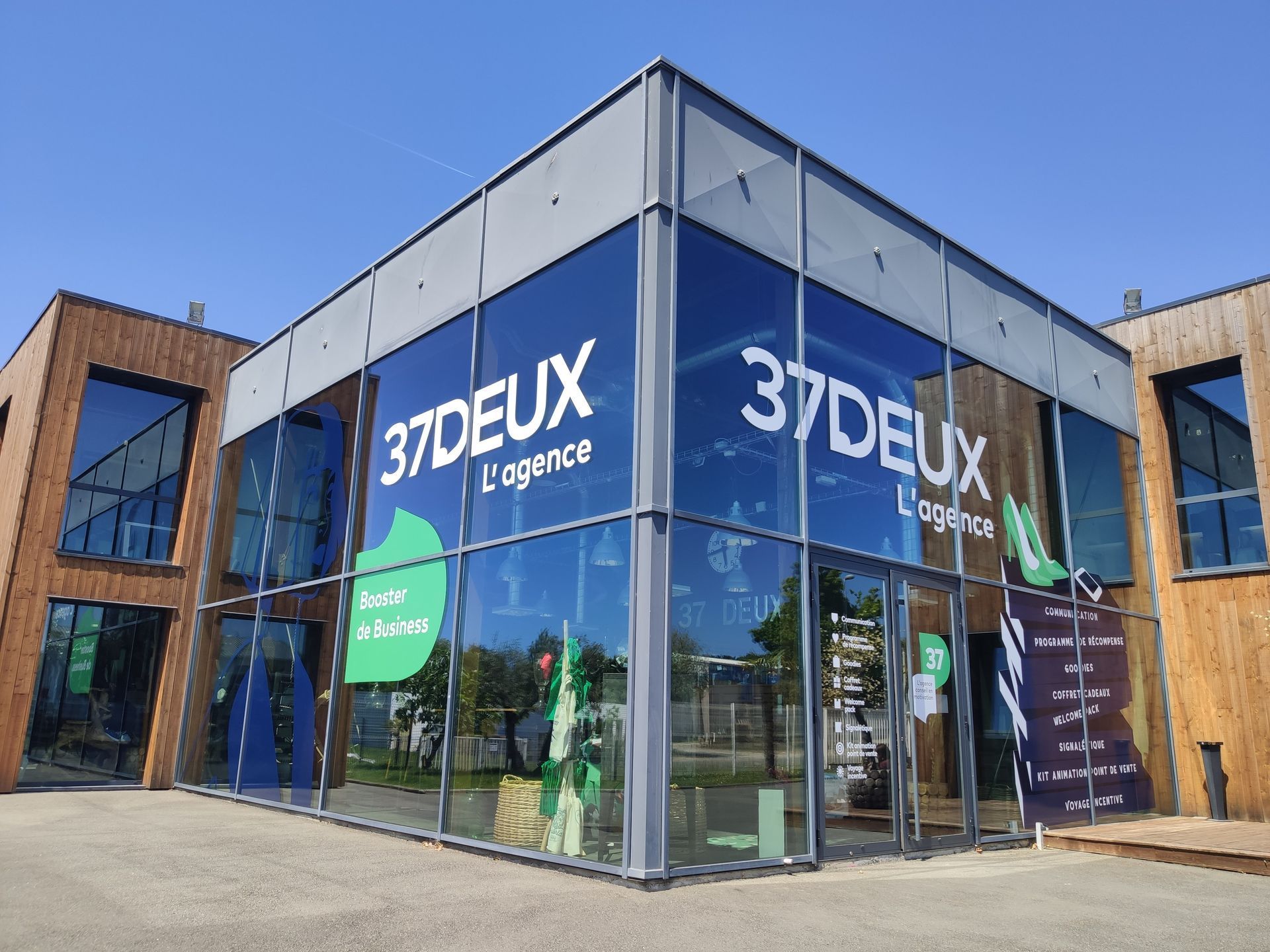 Vue d'angle d'un bâtiment moderne à façade vitrée arborant le logo 37DEUX L'agence sous un ciel bleu clair.