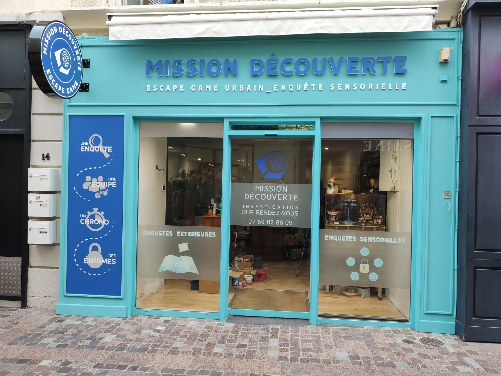 Façade de Mission Découverte, un établissement de jeux d'évasion de couleur turquoise situé dans une rue pavée.