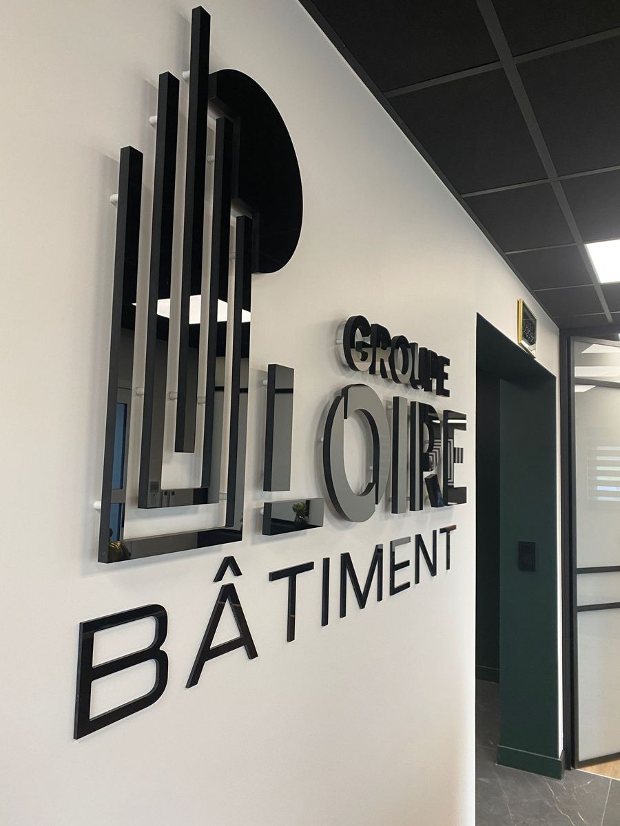 Une enseigne en 3D sur un mur blanc indique GROUPE LOIRE BÂTIMENT avec un grand dessin de bâtiment stylisé en noir.