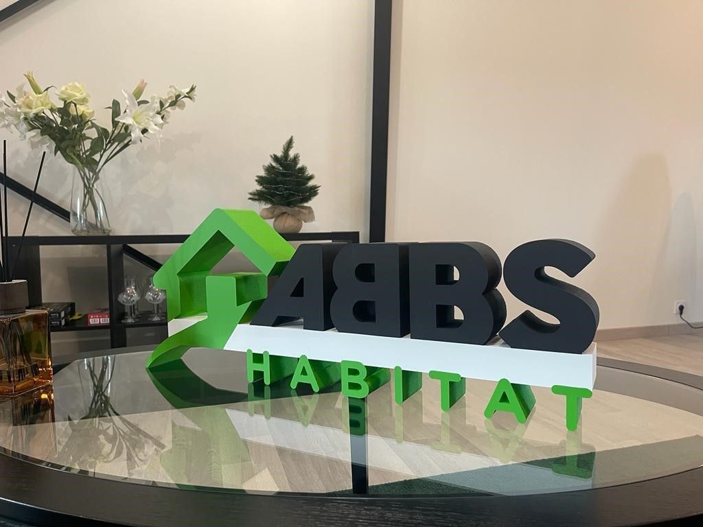 Un logo 3D vert et noir ABBS HABITAT est posé sur une table en verre dans une pièce moderne agrémentée d'une plante et de fleurs blanches.