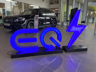 Un panneau EQ bleu lumineux et un symbole d'éclair sont exposés dans une salle d'exposition automobile, avec une Mercedes-Benz en arrière-plan.