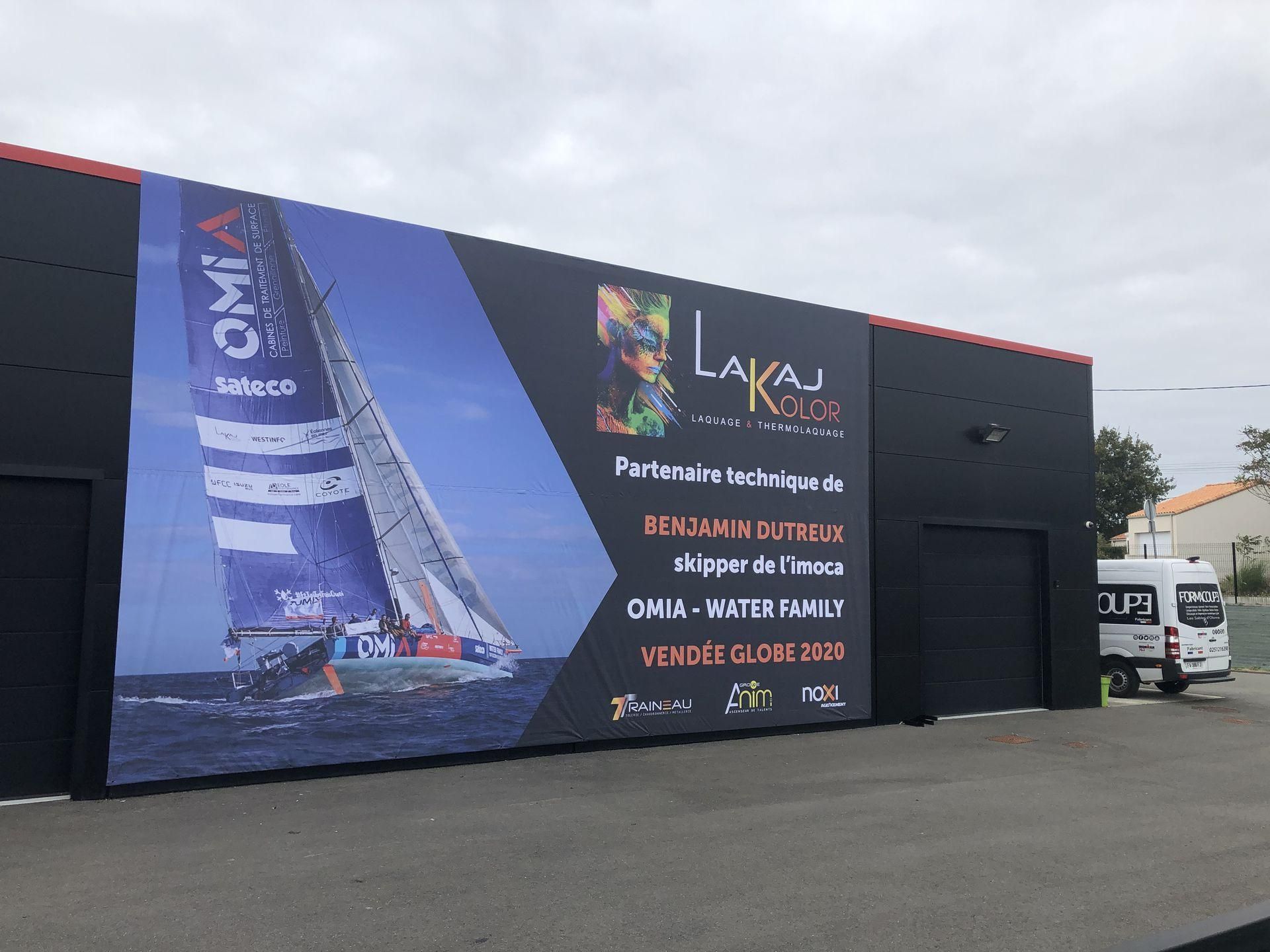 L'extérieur d'un bâtiment noir arbore un grand panneau publicitaire représentant un voilier en mer, avec les marques Lakaj Kolor et Omia.