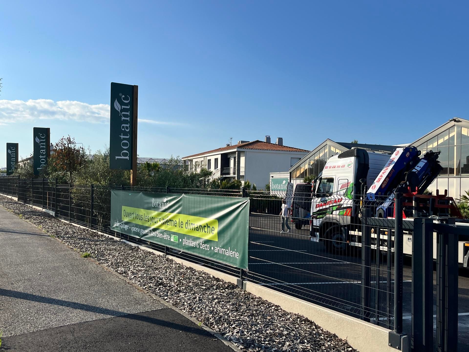 Un camion de livraison blanc est garé à côté d'une banderole verte portant le logo botanic, dans un espace extérieur sous un ciel dégagé.