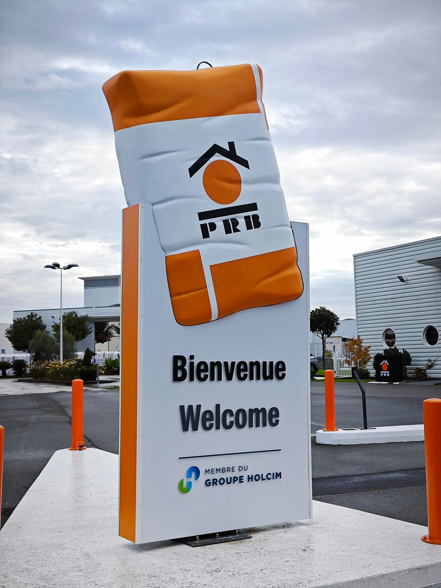 Un panneau de bienvenue blanc orné d'un sac orange portant le logo PRB, se dresse sur une zone pavée sous un ciel nuageux.