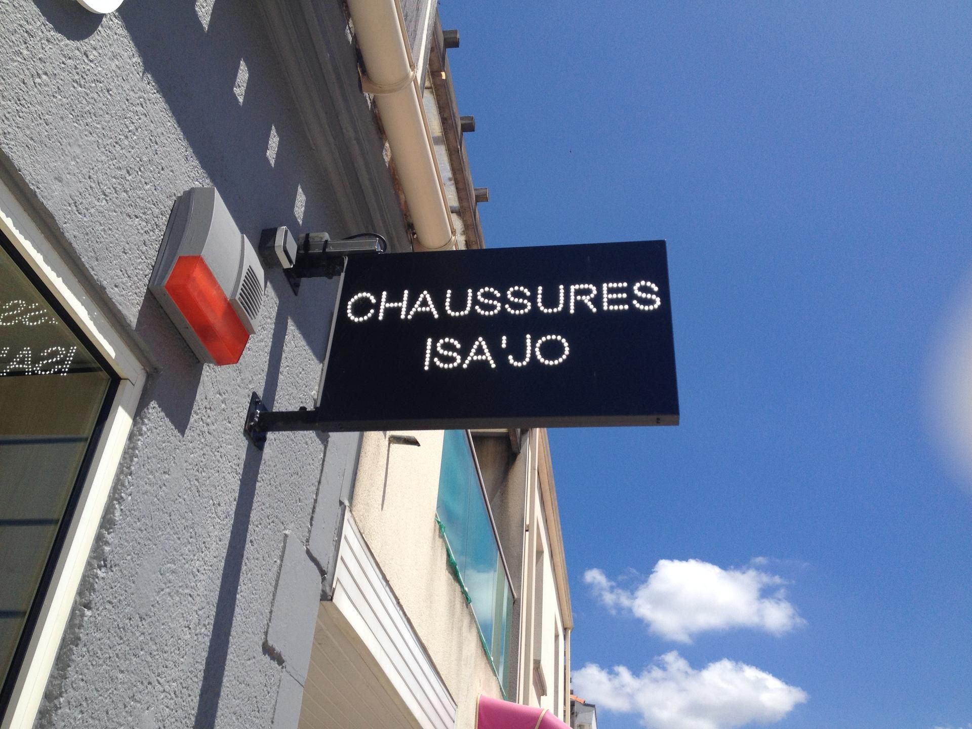 Une pancarte rectangulaire noire accrochée au mur gris d'un bâtiment, sur laquelle on peut lire CHAUSSURES ISA'JO en lettres capitales blanches.