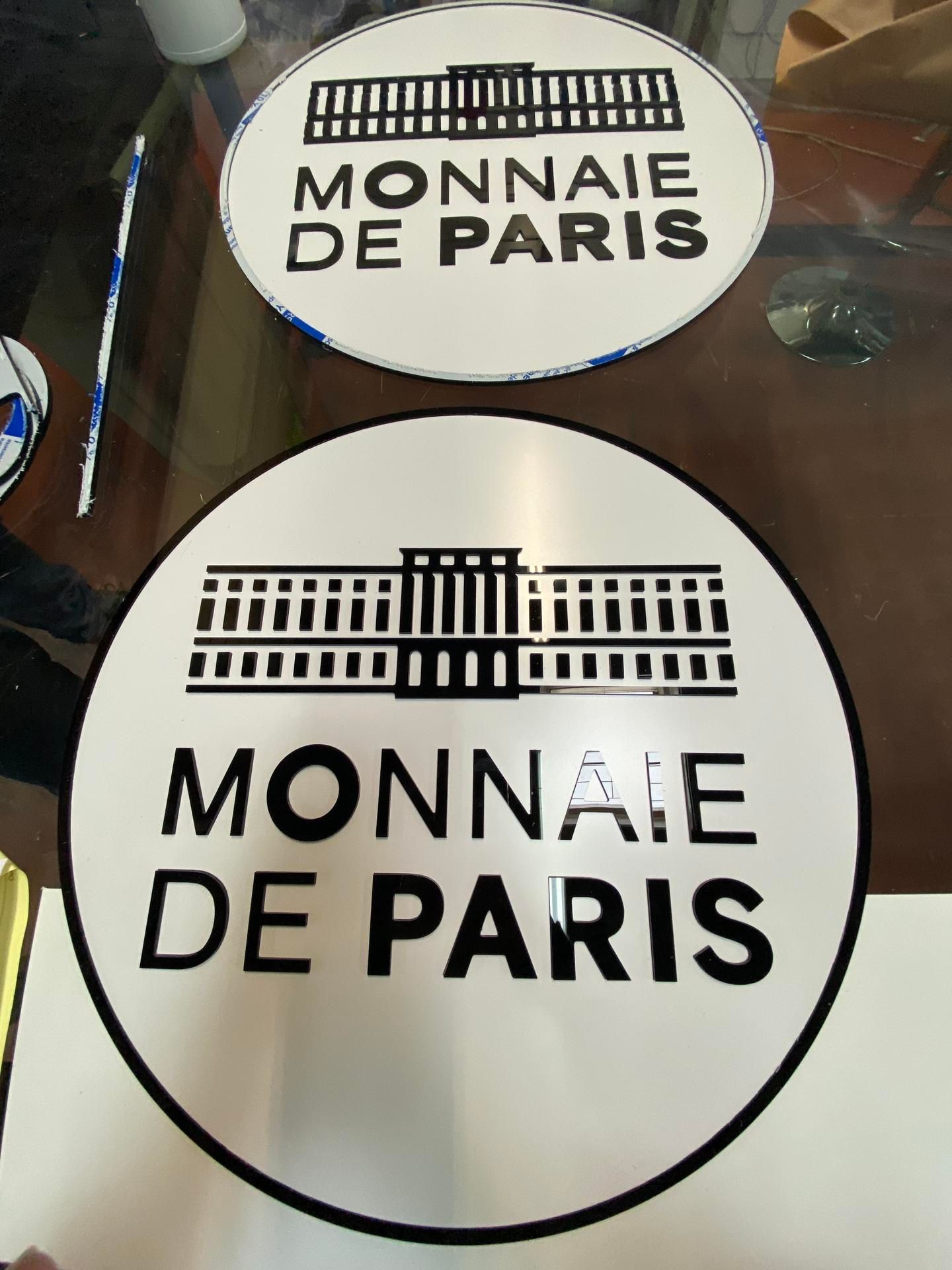 Deux panneaux circulaires blancs arborant le logo noir de la Monnaie de Paris, se détachant sur une surface intérieure floue.