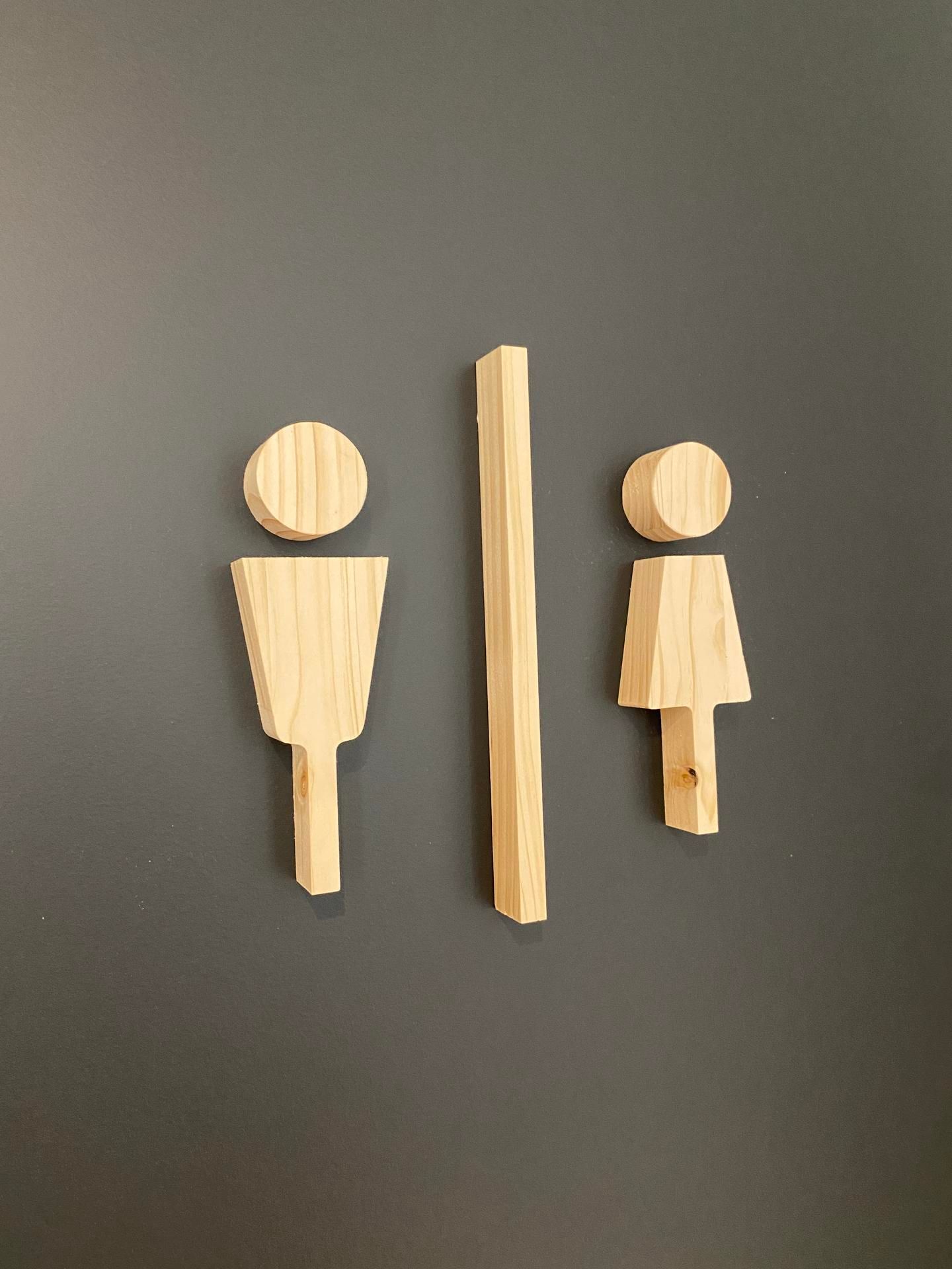 Panneau de toilettes comportant des icônes en bois clair représentant un homme et une femme, séparées par une barre verticale en bois, sur un mur gris.