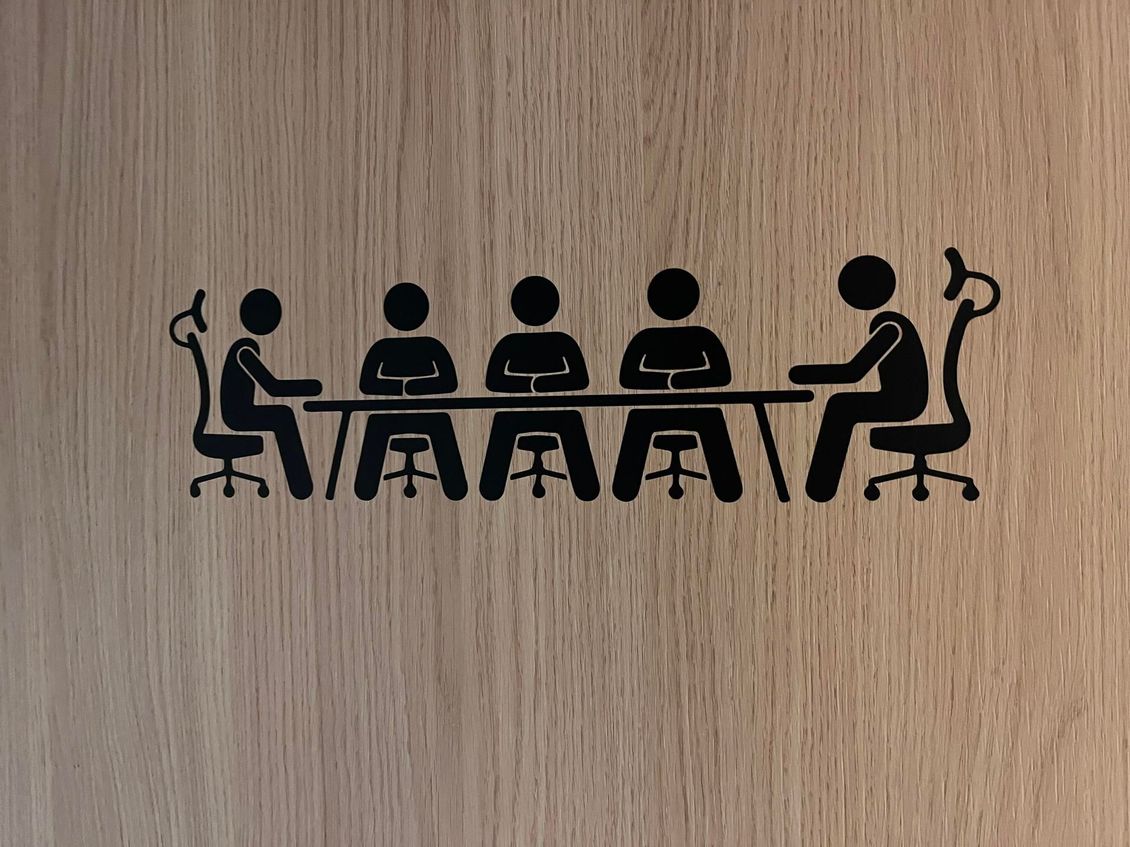 Une icône noire représentant cinq personnes assises autour d'une longue table de conférence, sur un fond en bois brun clair.