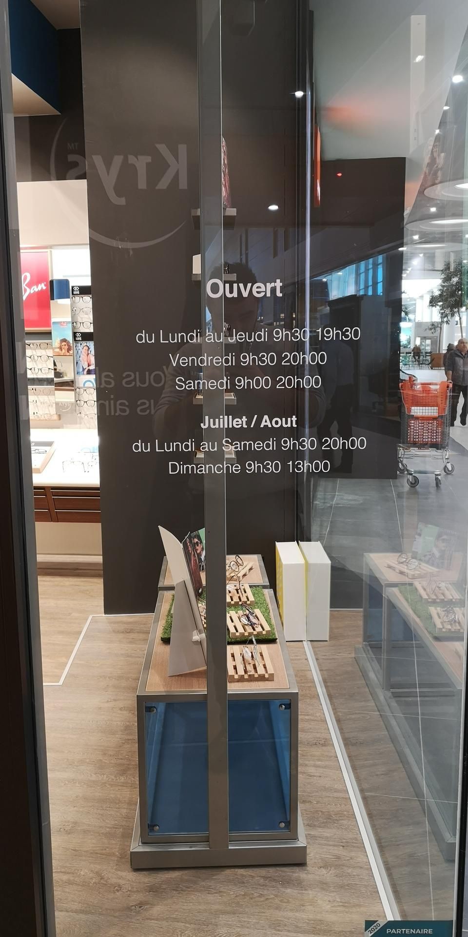 Porte vitrée d'un magasin d'optique avec les horaires d'ouverture imprimés en blanc, présentant des produits sur des étagères à l'intérieur.