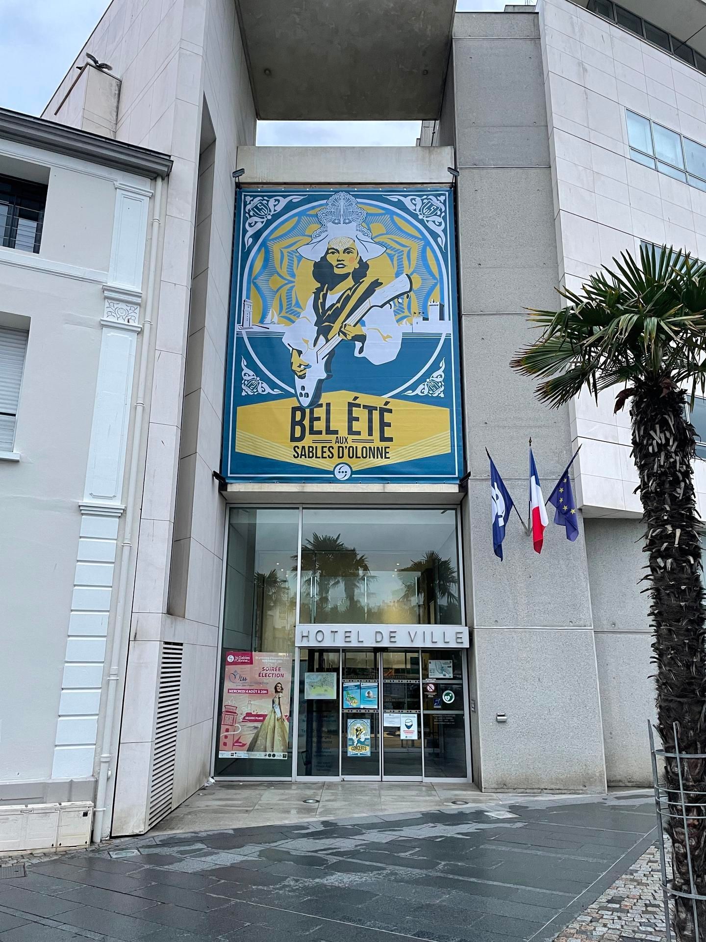 Entrée d'un hôtel de ville avec une grande affiche Bel été illustrée au-dessus des portes vitrées.