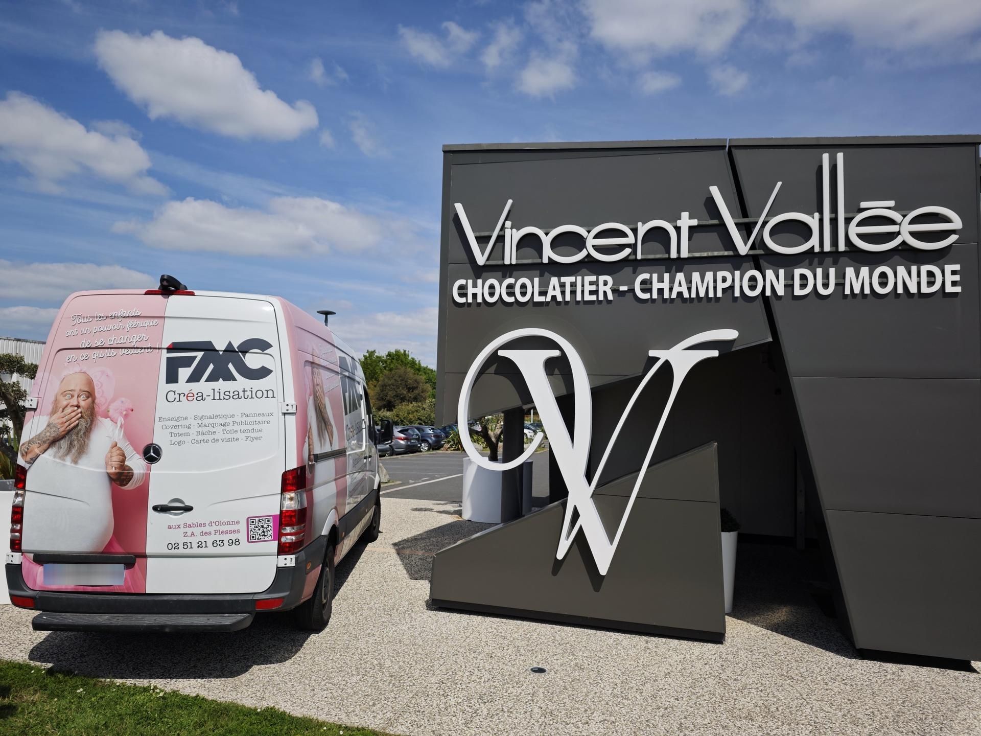 Une camionnette blanche ornée d'une publicité rose est garée à côté d'une enseigne moderne pour Vincent Vallée, Chocolatier, sous un ciel bleu.