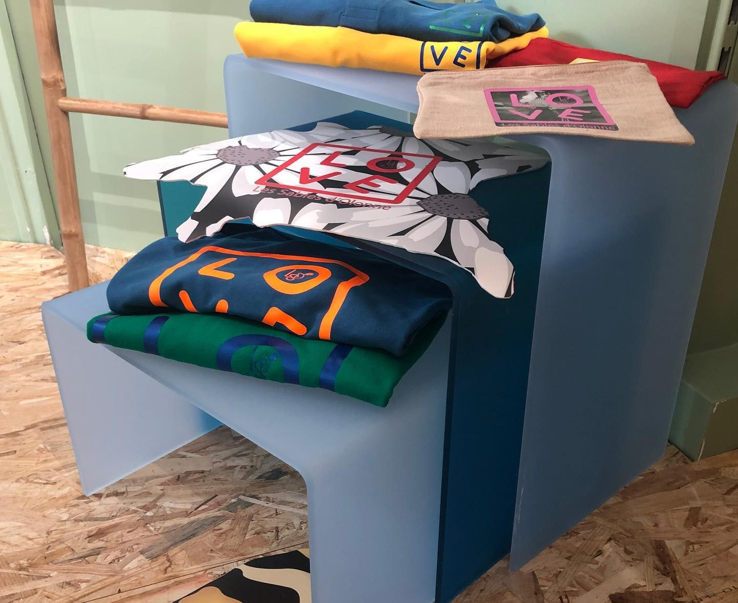 Des vêtements de marque colorés et pliés sont présentés sur un présentoir moderne bleu à plusieurs niveaux, au-dessus d'un sol en bois.