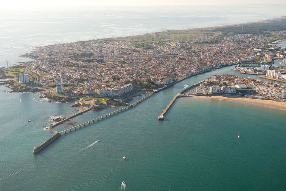 Vue aérienne d'une ville côtière montrant l'entrée de son port avec des brise-lames en pierre, des bâtiments et l'eau environnante.