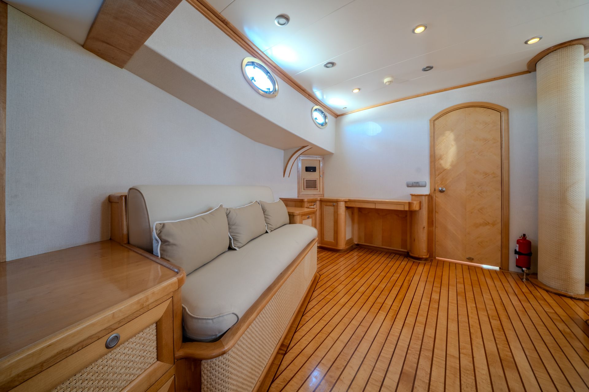 Un canapé beige trône dans une pièce minimaliste aux tons chauds du parquet, aux murs clairs, aux éclairages encastrés et à la porte en bois.