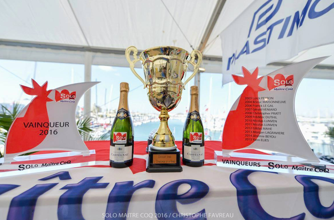 Un trophée en or trône entre deux bouteilles de champagne et deux récompenses en forme de coq sur une table ornée d'une bannière blanche et bleue.