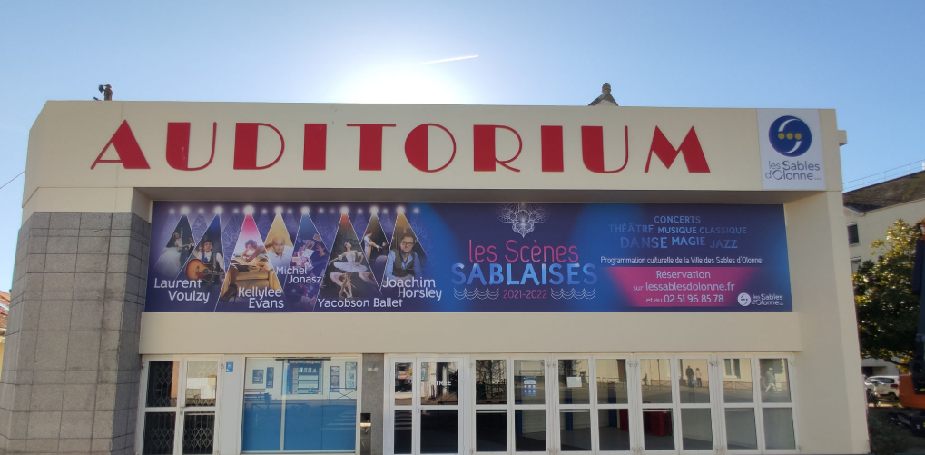 Extérieur d'un bâtiment abritant un auditorium, avec une grande banderole annonçant les Scènes Sablaises sur sa façade principale.