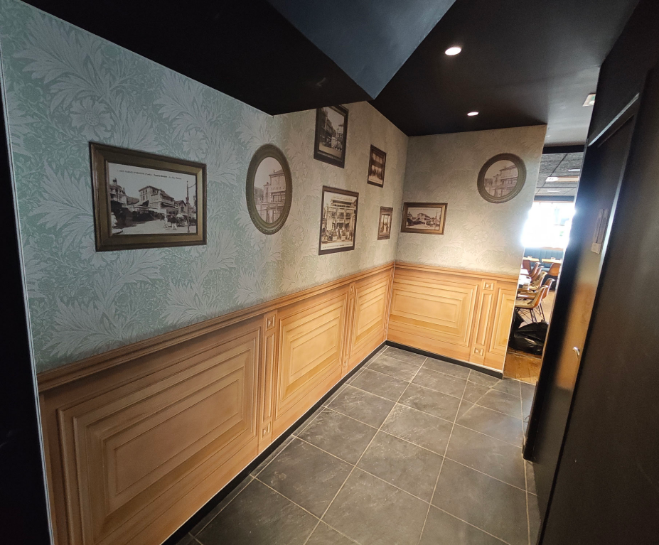 Un couloir intérieur avec du papier peint bleu clair à motifs, des lambris en bois et plusieurs tableaux encadrés aux murs.