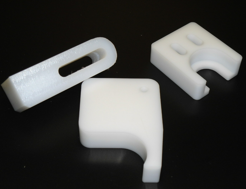 Trois composants en plastique blanc imprimés en 3D, de formes variées, dont une pièce à fente, sur fond noir.