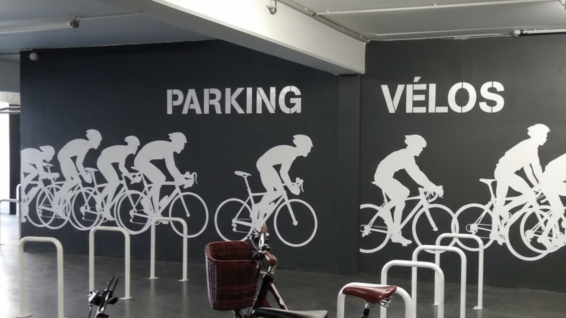 Parking à vélos intérieur avec des fresques murales représentant des cyclistes blancs et les mots PARKING et VÉLOS sur les murs sombres.