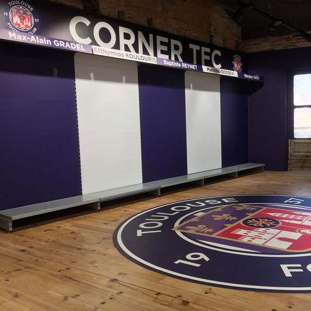 Vestiaire vide avec murs violets, sol en bois et enseigne CORNER TFC au-dessus des bancs.