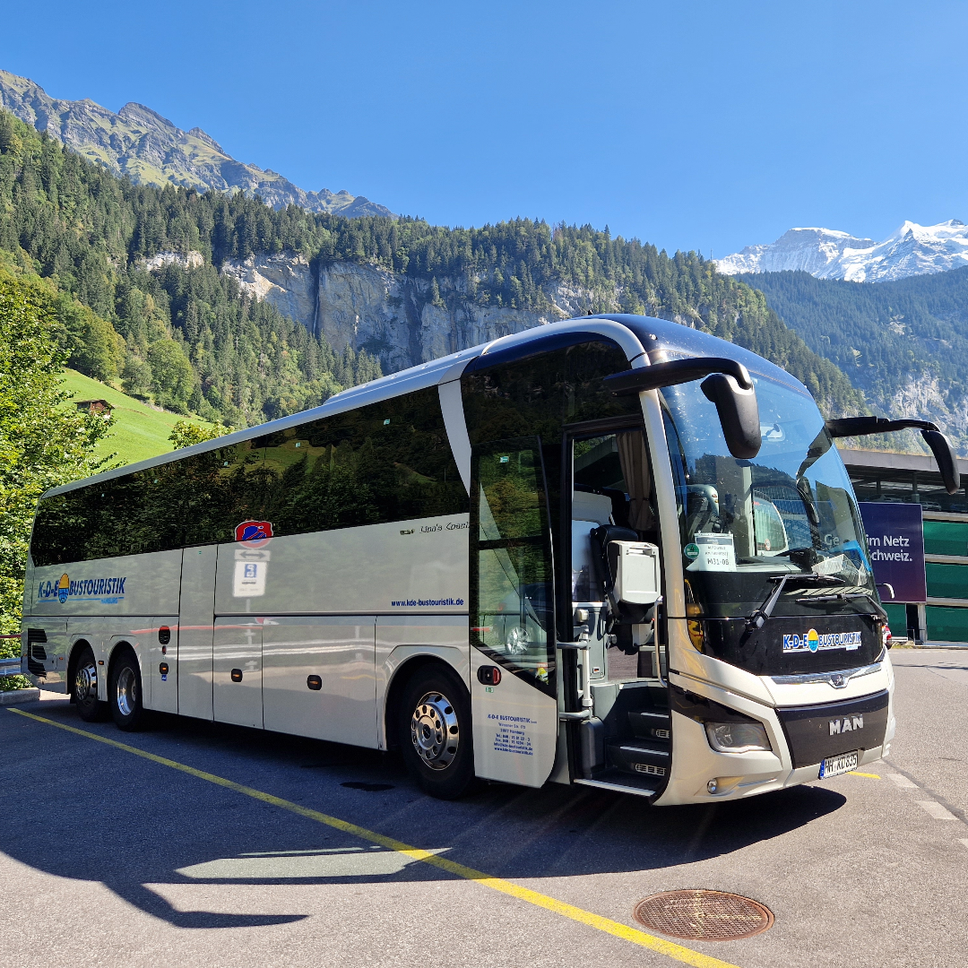 Ein cremefarbener Reisebus parkt mit offener Tür; im Hintergrund Berge. Sonniger Tag.