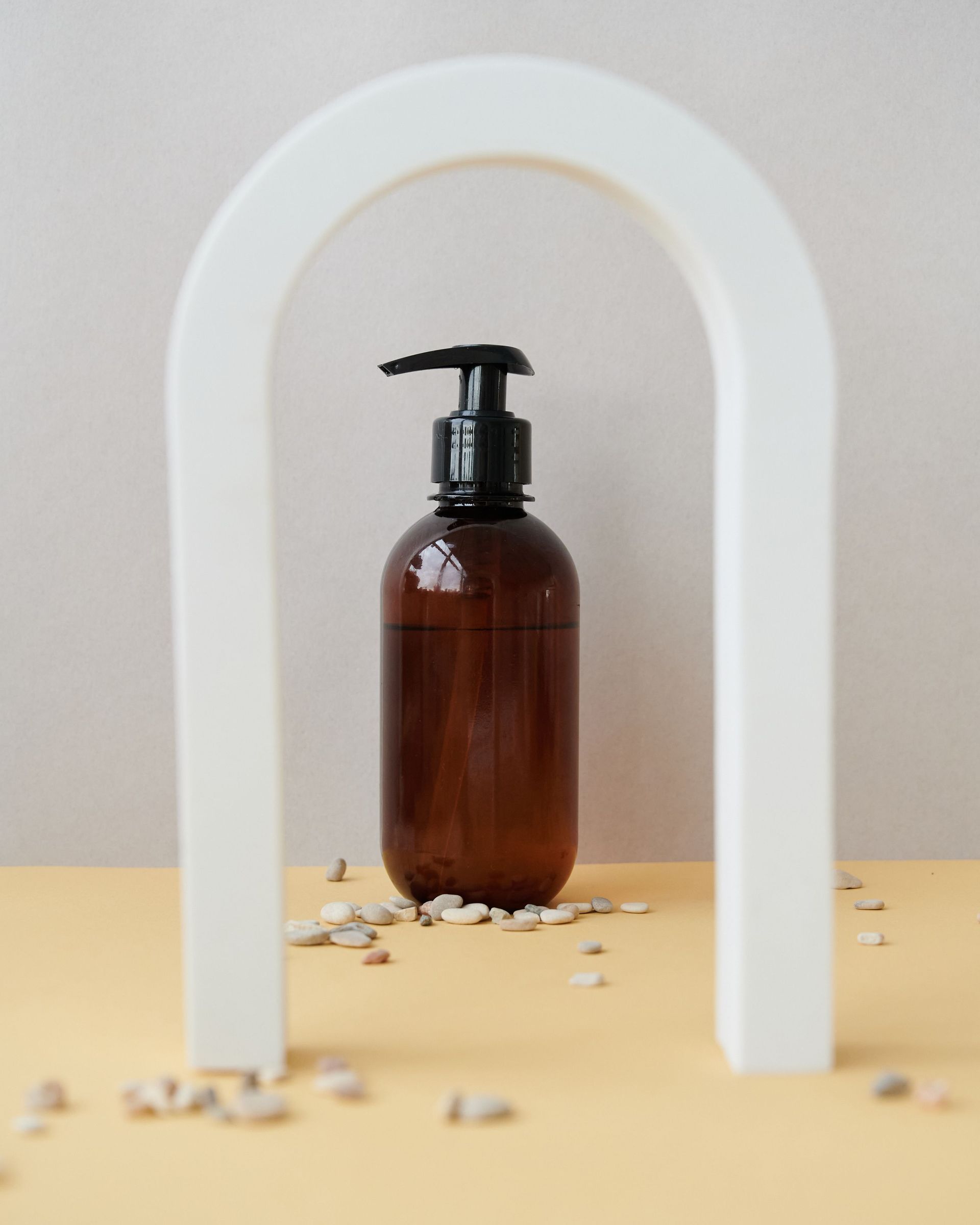 Un frasco de color ámbar con dosificador, enmarcado por un arco blanco, sobre una mesa beige con flores secas esparcidas.