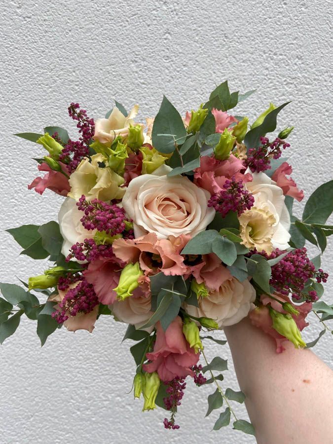 Ein handgebundener Blumenstrauß aus rosafarbenen, cremefarbenen und burgunderroten Blüten mit Eukalyptus vor einer grauen Wand