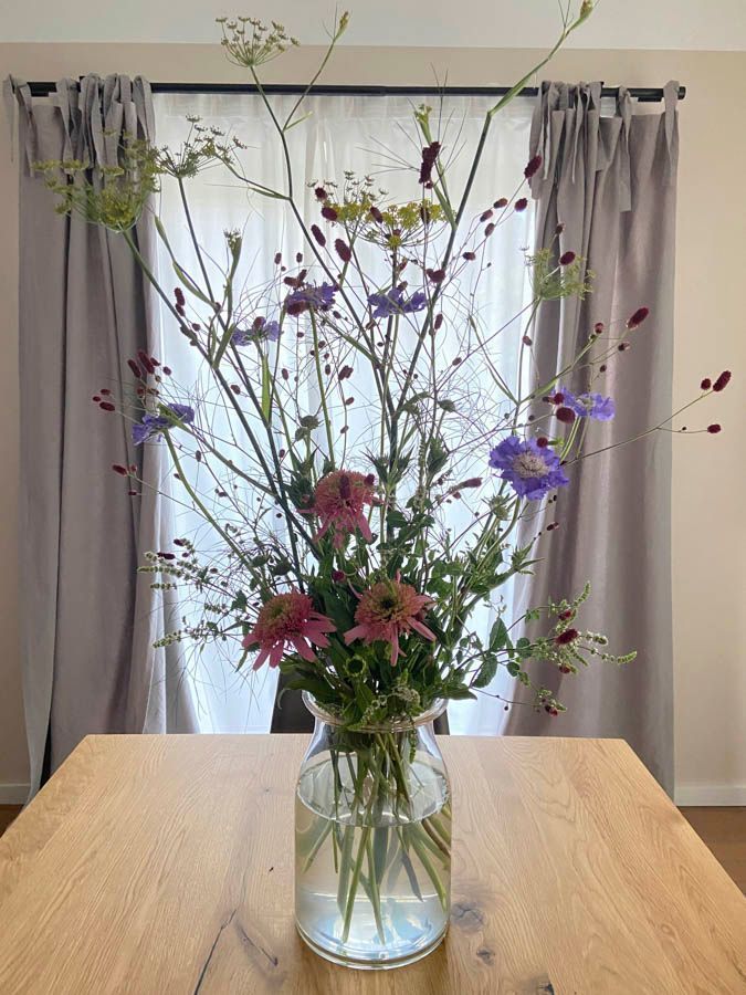 Ein hohes Wildblumenarrangement in einer Glasvase auf einem Holztisch vor verhängten Fenstern