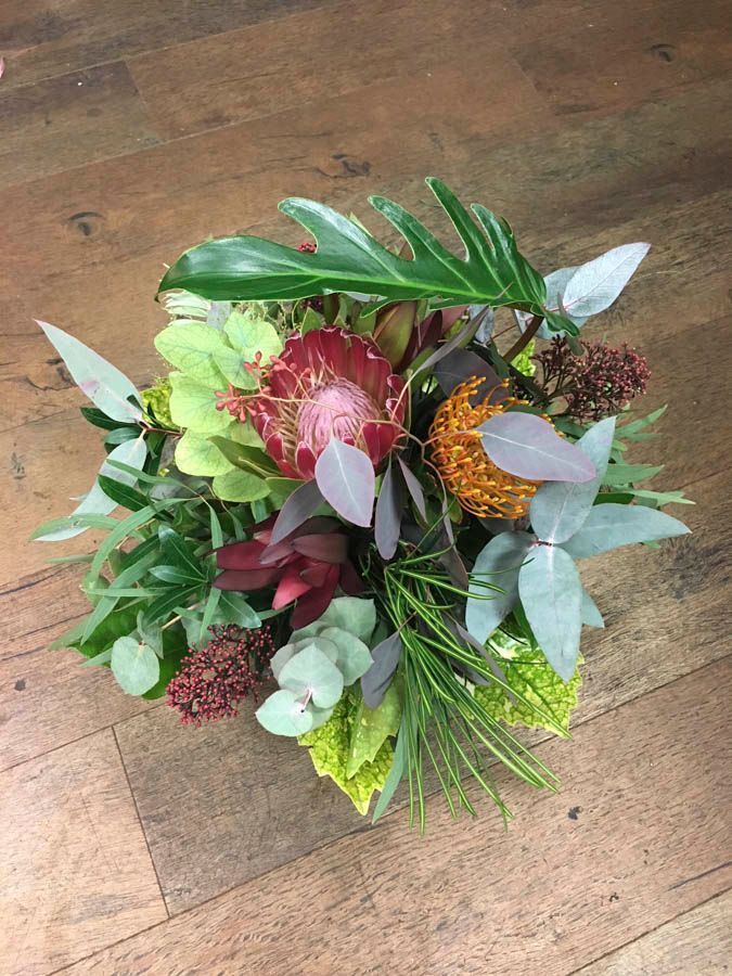 Tropisches Blumenarrangement mit grünen Blättern, rosa Blüten und orangefarbenen Akzenten auf einem Holzboden