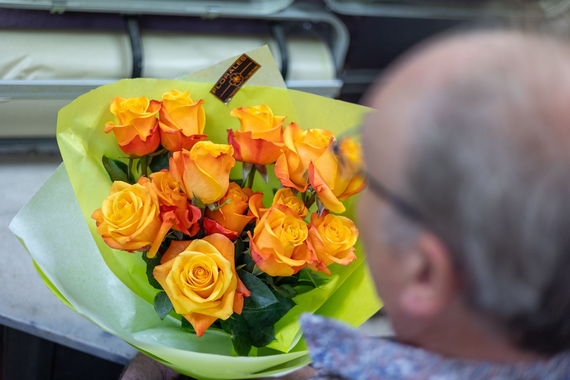 Ein Strauß orangefarbener Rosen, eingewickelt in hellgrünes Papier, wird vor einer verschwommenen, sitzenden Person gehalten.