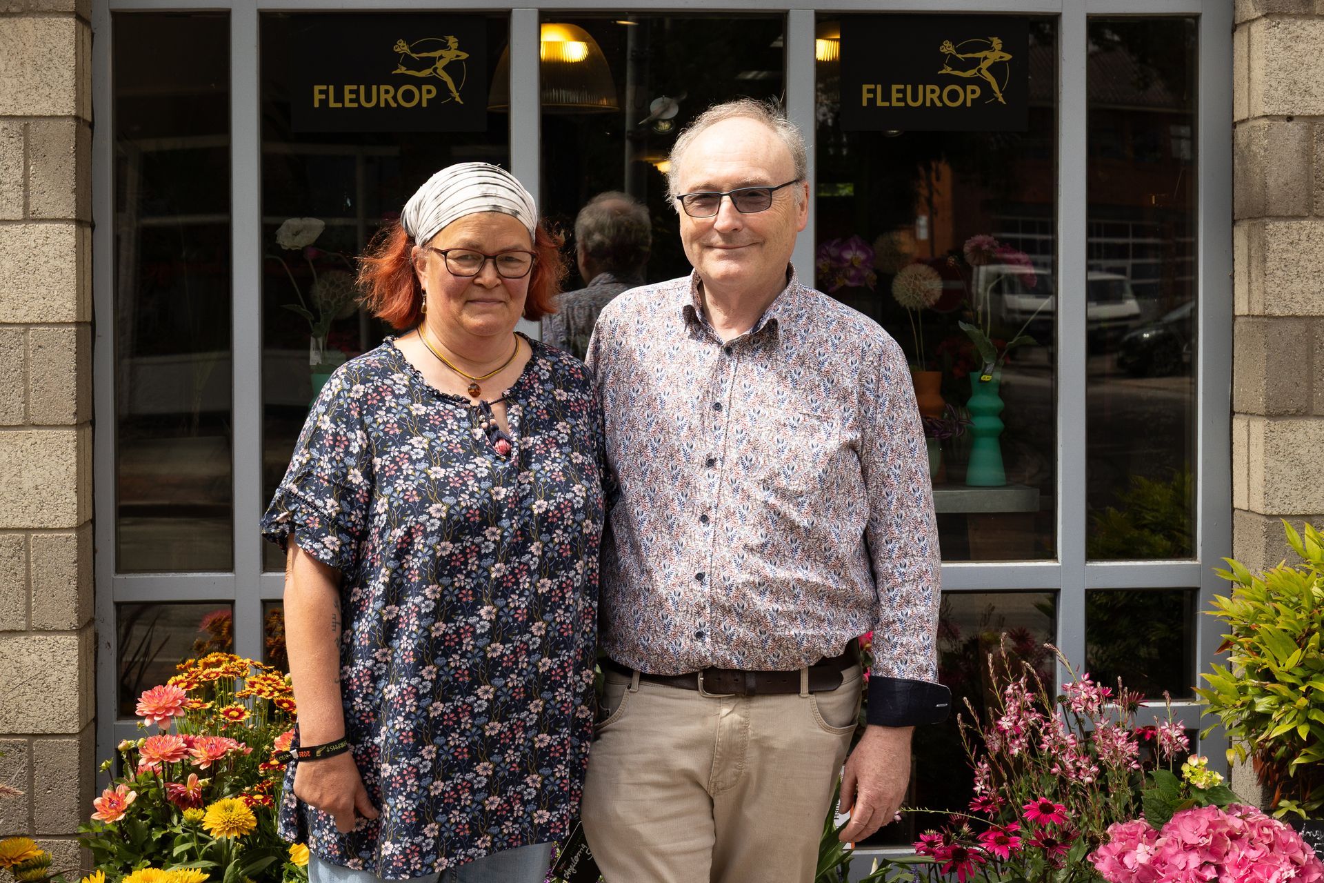 Zwei Personen stehen vor einem Blumenladen und lächeln vor den farbenprächtigen Blüten und der Schaufensterfront.