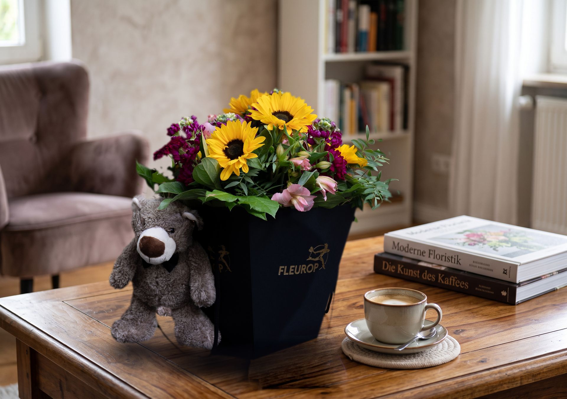 Ein Blumenarrangement in einem marineblauen Eimer auf einem Couchtisch, dazu ein Teddybär, Bücher und eine Tasse in einem gemütlichen Zimmer