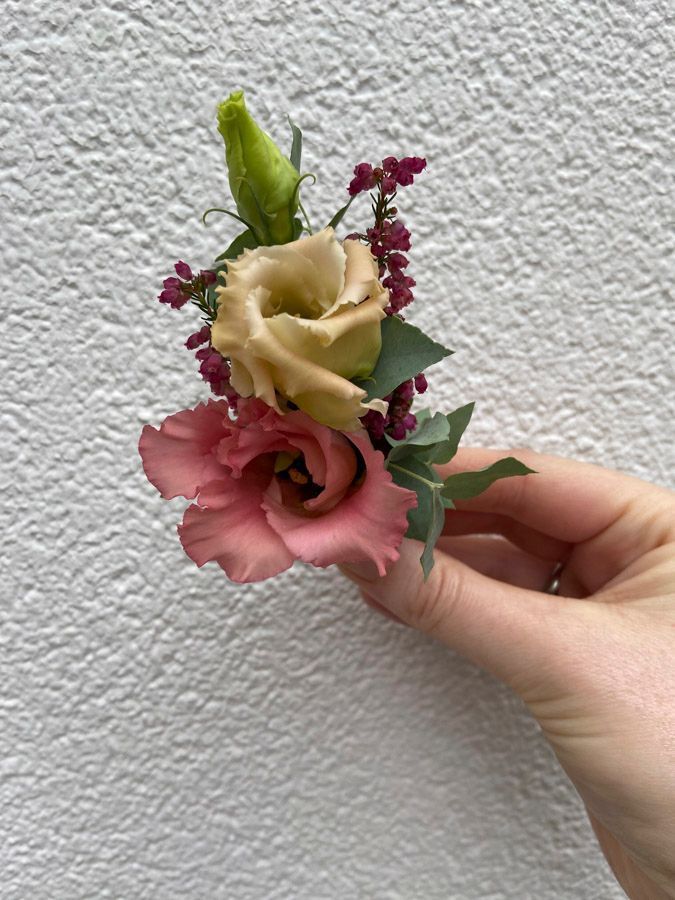 Eine Hand hält einen kleinen Strauß rosa und cremefarbener Blumen vor einer weißen Wand