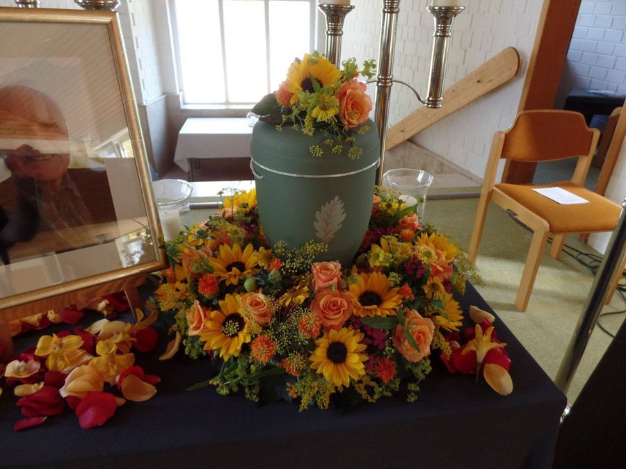 Blumengesteck als Gedenkarrangement mit gelben und orangen Blumen um eine Vase und einem gerahmten Porträt auf einem Tisch