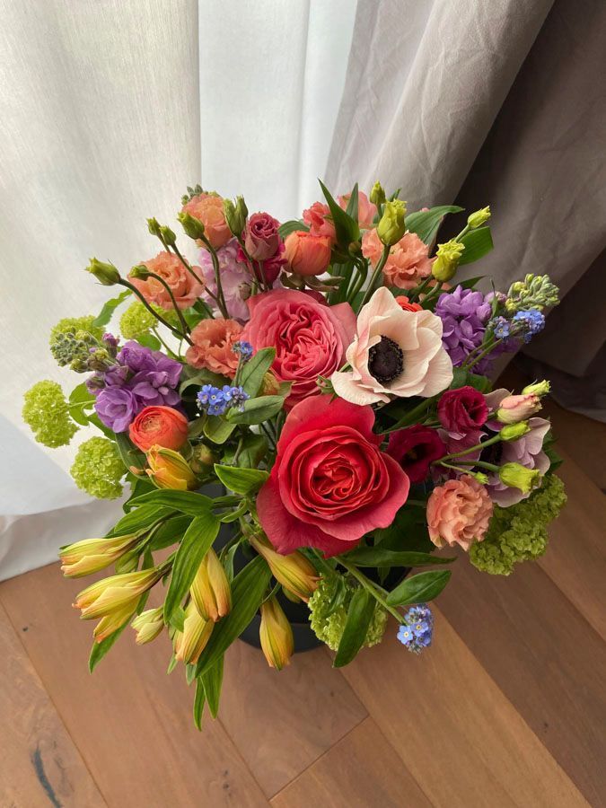Ein leuchtend bunter, gemischter Strauß aus Rosen, Lilien und lila Blumen in einer Vase auf einem Holzboden neben den Vorhängen.