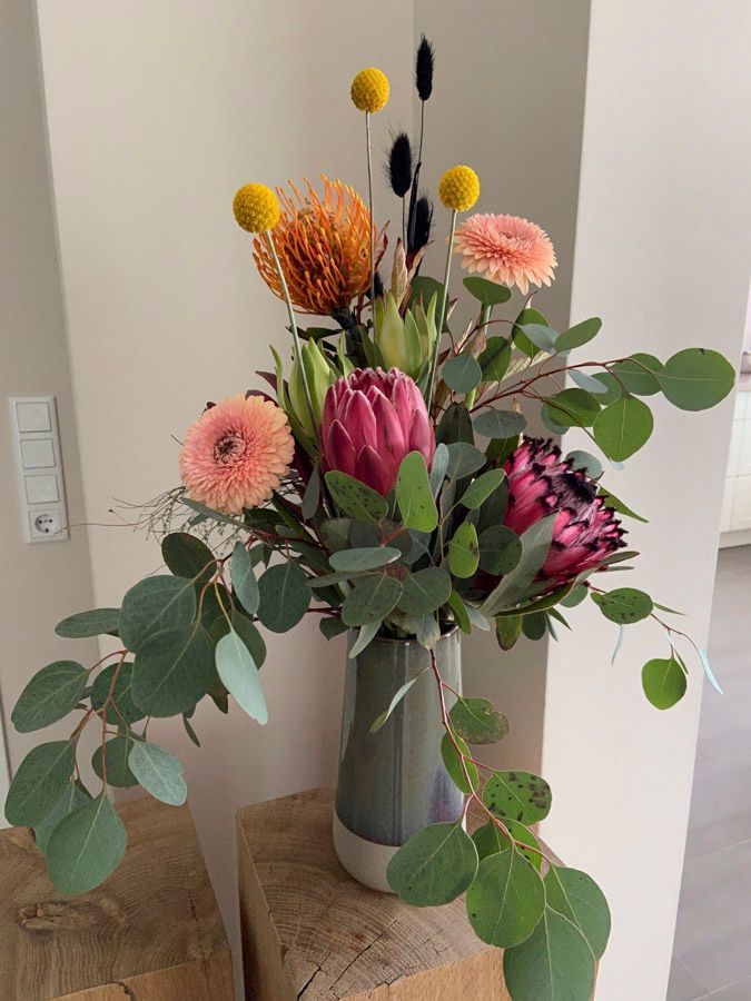 Ein bunter gemischter Blumenstrauß in einer grauen Vase auf einem Tisch, mit rosa, orangen, gelben und grünen Blüten.