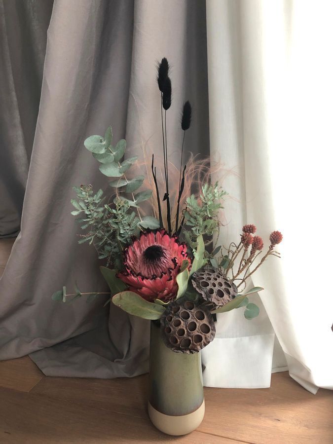 Rustikales Blumenarrangement in einer Vase mit rosa Protea, getrockneten Samenkapseln und Grünzeug an einem Vorhang