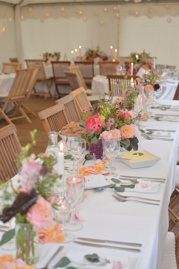 Eine lange Festtafel mit weißen Tischdecken, rosa Blumengestecken und Korbstühlen in einem Festzelt