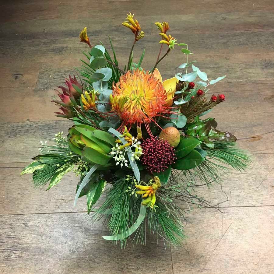 Farbenfrohes Blumenarrangement mit orangefarbenen, gelben, grünen und bordeauxroten Blüten auf einem Holzboden